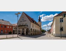Historisches Rathaus der Stadt Trebbin mit Arkadengang, einem dreieckigen Giebel und einem Wappen, daneben ein Kirchturm mit spitzem Dach; umgeben von weiteren Gebäuden in einer kleinen Stadtstraße unter klarem Himmel.