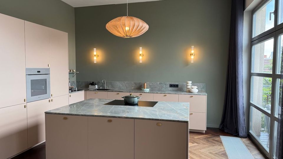 Modern kitchen with pale pink flat-panel cabinets and a large marble-look island featuring a black cooktop, set against a green wall with three wall sconces and a woven pendant light. Floor-to-ceiling windows with dark curtains bring in daylight above a wood herringbone floor and long floor vent.