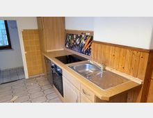 Kleine Küchenzeile mit Holzfronten, einer Spüle aus Edelstahl, einem eingebauten Kochfeld und einer Rückwand mit Gemüse-Motiv. Der Raum hat gelbe Wandfliesen, eine Holzverkleidung an der Wand und einen Boden mit beigen Fliesen in Sechseckform.