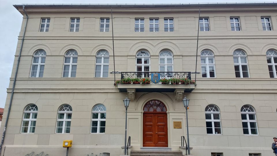 Fassade eines historischen Gebäudes mit beigen Wänden, weißen Fenstern im Rundbogenstil und einem großen Holzeingangstor. Über dem Eingang befindet sich ein kleiner Balkon mit Blumen und einem Wappen, flankiert von zwei Laternen.
