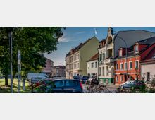 Straßenszene in einer Kleinstadt mit mehrstöckigen Wohngebäuden in verschiedenen Farben, darunter ein auffälliges Haus mit grünen Verzierungen und Bogenfenstern. Im Vordergrund sind geparkte Autos, ein Parkplatzschild und Bäume zu sehen.