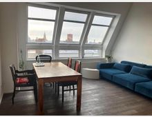 Attic-style living and dining room with a sloped ceiling and large angled skylight windows overlooking city rooftops. A wooden dining table with chairs sits on dark flooring, with a blue sectional sofa and small white side tables along the wall.