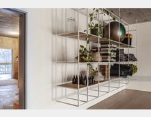 Ein minimalistischer Innenraum mit einem weißen, offenen Regal aus Metall. Im Regal befinden sich Pflanzen, Glasgefäße, gefaltete Handtücher, Yogamatten, Fitnesszubehör und kleine braune Flaschen. Durch eine Tür ist ein Raum mit Holzverkleidung und großem Fenster sichtbar.