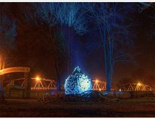 Ein nächtliches Bild zeigt eine beleuchtete Skulptur aus Holzästen in einer blauen Lichtinstallation, umgeben von kahlen Bäumen. Im Hintergrund sind eine beleuchtete Brücke und ein Schild mit der Aufschrift „Willkommen am …“ sichtbar.