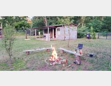 Ein einfacher Garten mit einer Feuerstelle im Vordergrund, umgeben von Holzbänken, und einer kleinen Hütte im Hintergrund. Der Garten ist von Bäumen und Büschen umgeben, und eine Person steht spielend auf der Wiese.