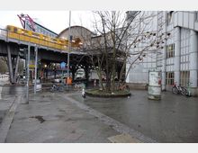 Straßenszene in einer Stadt mit einer erhöhten Gelbbahn auf einer Stahlkonstruktion, daneben ein modernes Gebäude mit Glas- und Gitterelementen. Im Vordergrund befinden sich eine Fußgängerzone mit nassem Pflaster, Bäumen, Fahrrädern und E-Scootern sowie eine Litfaßsäule.