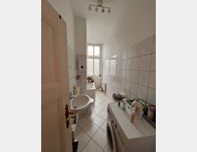 Schmales Badezimmer mit weiß gefliesten Wänden und Boden. Zu sehen sind ein Waschbecken, eine Badewanne mit Fenster darüber, ein Waschmaschine im Vordergrund und verschiedene Hygieneartikel auf Ablagen.