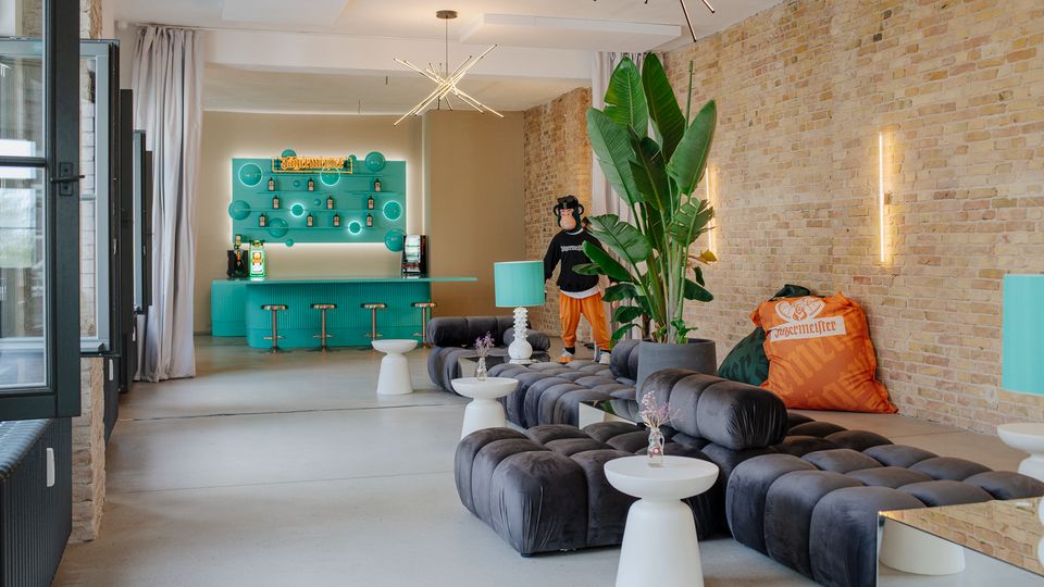 Moderner Lounge- und Barbereich mit freiliegender Ziegelwand, hohen Decken und türkisfarbener Theke samt Regalen im Hintergrund. Im Vordergrund stehen modulare, dunkelgraue Polstersofas mit kleinen weißen Beistelltischen, dazu große Zimmerpflanzen und filigrane Deckenleuchten.