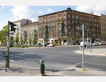 Straßenkreuzung mit mehrstöckigem Altbau im Vordergrund, der mit roten Ziegeln und detaillierten Fassadenelementen gestaltet ist. Im Erdgeschoss befinden sich kleine Geschäfte und Cafés, während parkende Autos und eine Ampel den Verkehr markieren.