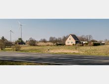 Ländliche Landschaft mit einem Einfamilienhaus und mehreren Nebengebäuden auf einer grünen Wiese, im Hintergrund zwei Windkraftanlagen und vereinzelt stehende Bäume. Im Vordergrund verläuft eine asphaltierte Landstraße.