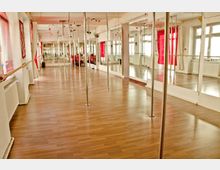 Ein heller Trainingsraum mit Tanzstangen, großen Wandspiegeln und einem Holzboden. Rote Vorhänge und Fenster sorgen für zusätzliches Licht und Akzente.