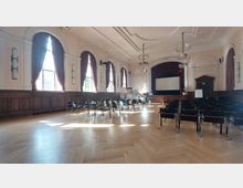 Ein heller Veranstaltungssaal mit hohen Decken, großen Fenstern mit weinroten Vorhängen und einem Holzboden in Fischgrätoptik. Vorne befindet sich eine Bühne mit Leinwand sowie aufgereihte Stuhlreihen im Raum.