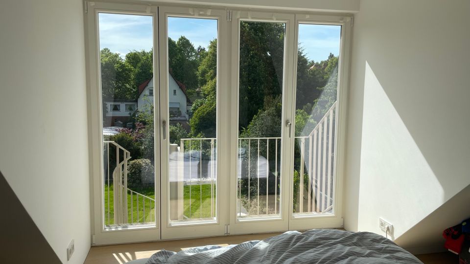 Helles, schlichtes Schlafzimmer mit großen bodentiefen Fenstertüren und kleinem Balkon mit weißem Geländer. Durch die Fenster blickt man auf einen grünen Garten mit Bäumen und benachbarte Häuser.