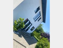 Schräg aufgenommene Außenansicht eines modernen weißen Wohnhauses mit großen rechteckigen Fenstern und dunklem Dachüberstand. Davor liegt eine gepflasterte Einfahrt mit kleinen Büschen und Bäumen sowie angrenzendem Grünbereich.