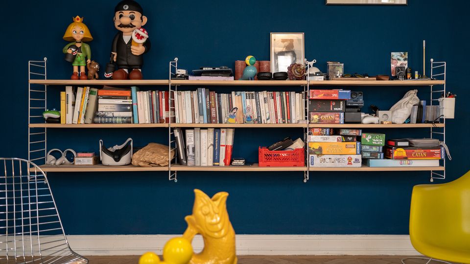 Wall-mounted shelving on a dark blue living-room wall filled with books, board games, and small decorative objects, with framed artwork above. Modern seating includes a wire chair and a yellow chair, and a low table holds a yellow sculptural piece in the foreground.