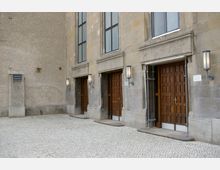 Steinfassade eines Gebäudes mit drei großen Holztüren und hohen rechteckigen Fenstern darüber, flankiert von metallenen Wandlampen. Der Boden besteht aus einem gepflasterten Hof mit hellem Steinbelag.