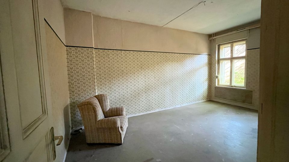 Leerer Innenraum eines unsanierten Zimmers mit grauem Boden, gemusterter Tapete und hohen Decken. Rechts befindet sich ein großes Sprossenfenster über einem Heizkörper; links ist eine Tür und ein einzelner Sessel steht in der Ecke.