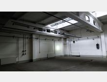 Leerer, industrieller Innenraum mit weiß gestrichenen Wänden und sichtbaren Rohren. Die Decke hat Oberlichter und hängende Leuchtstofflampen, der Boden besteht aus glattem Beton.