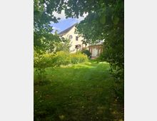 Ein Garten mit grünem Rasen und Bäumen, im Hintergrund ein weißes Gebäude mit schrägem Dach, einem runden Fenster und einem Balkon. Sträucher und gelb blühende Pflanzen umgeben das Gebäude.