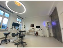 Moderne Zahnarztpraxis mit einem weißen Behandlungsstuhl, umgeben von zahnmedizinischem Equipment, mehreren Bildschirmen und einem runden Deckenlicht. Der Raum hat helle Wände, große Fenster mit Tageslicht und ein Röntgengerät an der rechten Wand.