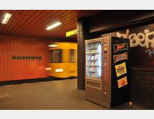 U-Bahnhof mit orangefarbener Wand mit der Aufschrift „Halemweg“, einem einfahrenden gelben Zug auf der linken Seite und einem Snackautomaten auf der rechten Seite vor einer dunklen Wand mit Graffiti.