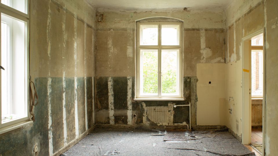 Leerer, entkernter Innenraum mit abgenutzten, teils abgeplatzten Wänden und beschädigtem Bodenbelag. Zwei hohe Fenster und ein Türdurchgang lassen Tageslicht herein; unter dem mittleren Fenster sind alte Heizungsrohre sichtbar.