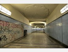 Ein unterirdischer Fußgängertunnel mit glatten, hellblauen Fliesenwänden, grauem Bodenbelag und abgerundeten Deckenbögen. Die Wände sind teilweise mit Graffiti bedeckt, und an der Decke befinden sich eingelassene Deckenlampen, die den Bereich beleuchten.