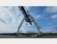 Großer Hafenkran mit A-förmigem Stahlrahmen, Seilzug und Plattformen, stehend auf einer betonierten Fläche vor einer Reihe von Bäumen unter leicht bewölktem Himmel.