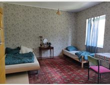 Ein einfach eingerichtetes Schlafzimmer mit zwei Einzelbetten, blauer Bettwäsche und Blumendekor an den Tapeten. Ein kleines Fenster mit blauen Vorhängen, ein Tisch mit kariertem Tuch, ein Stuhl und ein Teppich mit orientalischem Muster prägen den Raum.