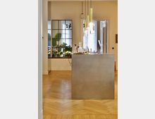 Innenansicht eines modernen Wohnraums mit Holzboden im Fischgrätmuster, einem schlanken, metallischen Küchentresen und hängenden, goldfarbenen Lampen. Im Hintergrund sind große Fenster, eine Glaswand mit schwarzen Streben und grüne Zimmerpflanzen zu sehen.