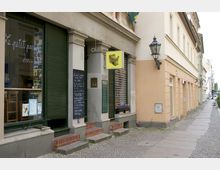 Straßenszene mit einem kleinen Geschäft mit grünen Rollläden und einer beschrifteten Tafel neben dem Eingang. Das Gebäude ist beige, mit typischer Altbauarchitektur, inklusive eines Wandlaternenleuchters.