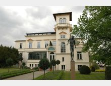 Eine historische Villa mit heller Fassade, großem Turm und bodentiefen Fenstern. Im Vordergrund befindet sich eine Statue auf einem hohen Sockel, umgeben von einer gepflegten Gartenanlage mit Kiesweg und Bäumen.