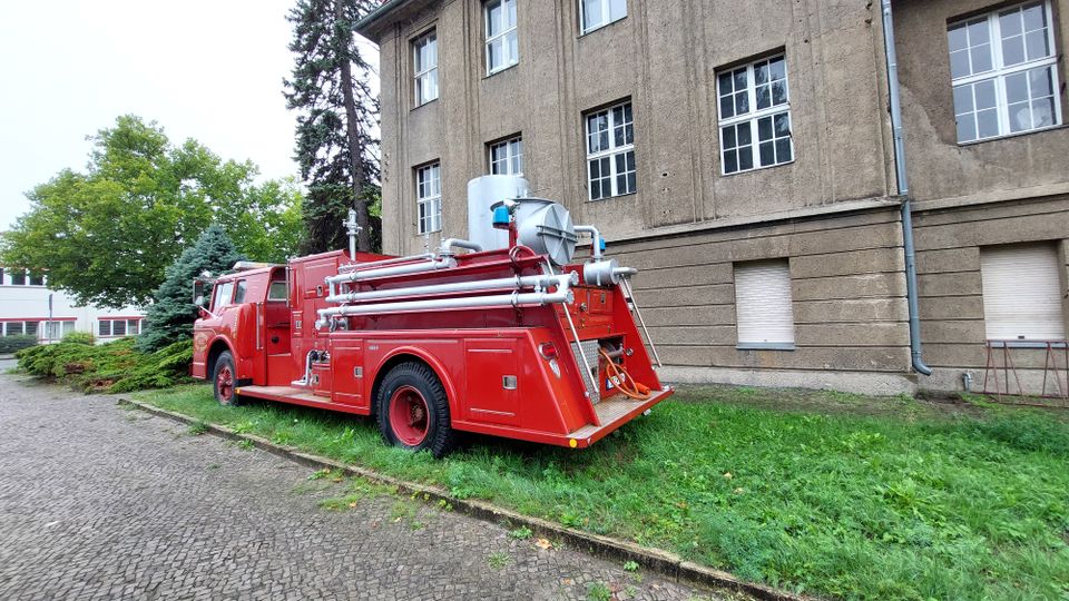 Ein rotes, historisches Feuerwehrfahrzeug mit Schläuchen und Metallaufbauten steht auf einer Rasenfläche neben einem großen, beige verputzten Gebäude mit mehreren Fenstern. Im Hintergrund sind Bäume und ein weiteres Gebäude zu sehen.
