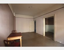 Ein einfacher, leerer Raum mit hellen Wänden und einem grauen Fußboden. Im Vordergrund stehen ein schlichter Holztisch und zwei Holzstühle, während im Hintergrund eine geschlossene weiße Tür und ein Durchgang zu einem angrenzenden Raum zu sehen sind.