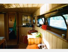 Holzvertäfelter Innenraum eines Hausboots mit schmalen Fenstern auf der rechten Seite, durch die Wasser und Bäume sichtbar sind. Im Vordergrund stehen ein kleiner Holztisch, eine Kommode und diverse Alltagsgegenstände, während im Hintergrund ein Durchgang zu einem weiteren Raum führt.