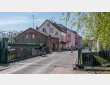 Straßenszene in einer kleinen Stadt mit Kopfsteinpflaster, einer Brücke aus Metallgeländern und Backsteinhäusern. Ein Radfahrer überquert die Brücke, flankiert von Gebäuden und Bäumen im Hintergrund.
