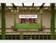 U-Bahn-Station "Deutsche Oper" mit grünen Metallstützen, einem grünen Sitzbankbereich und einem Schild mit der Aufschrift „Deutsche Oper“. Im Hintergrund eine bunte Wandgestaltung mit abstrakten Mustern.