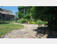 Ein ländlicher Garten mit einem gepflasterten Steinweg, der von Pflanzen und Blumen in bunten Farben gesäumt ist. Im Hintergrund ist ein Haus mit einer begrünten Fassade sowie ein altes Backsteingebäude zu sehen, während Bäume und Rasenflächen die Umgebung prägen.