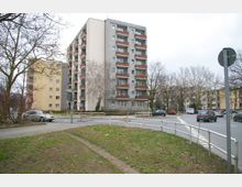 Mehrstöckiges Wohngebäude mit grau-roter Fassade und Balkonen, umgeben von weiteren Wohnhäusern. Im Vordergrund eine Straße mit parkenden Autos, ein kleiner Grünstreifen und kahler Baumbestand.