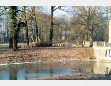 Ein herbstlicher Park mit kahlen Bäumen, einer leichten Laubdecke auf dem Boden und einem kleinen, teilweise zugefrorenen Teich im Vordergrund. Rechts im Bild befinden sich Steingeländer und Balustraden, die zu einem historischen Gebäude gehören könnten.