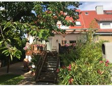 Zweistöckiges Wohnhaus mit rotem Ziegeldach und einem Holzbalkon, geschmückt mit bunten Blumen. Im Vordergrund befinden sich ein Garten mit Bäumen, Sträuchern und einer Treppe, die zum Balkon führt.