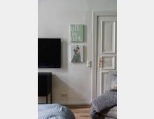 Ein modern eingerichtetes Wohnzimmer mit weißer Wand, an der zwei gerahmte Bilder hängen, eines mit dem Text „all is art“ und das andere mit einer Figur in grünem Kleid. Rechts daneben eine weiße Tür mit klassischen Holzvertäfelungen, links ein schwarzer Flachbildfernseher, und im Vordergrund ein graues Sofa mit Decke.