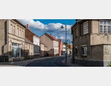 Straßenszene in einer Kleinstadt mit zweistöckigen Häusern auf beiden Seiten, größtenteils mit verputzten Fassaden und vereinzelten dekorativen Elementen. Die schmale, asphaltierte Straße ist von Bürgersteigen gesäumt, und historische Straßenlaternen sind entlang der Fahrbahn aufgestellt.