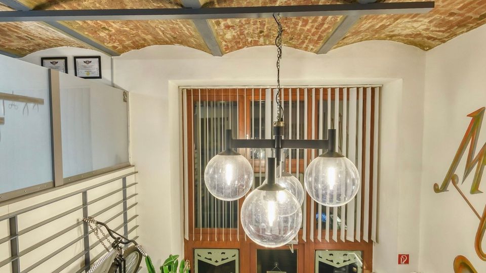 Blick von einer Empore in einen kleinen Laden mit freiliegender Ziegeltonnen-Decke und dunklen Stahlträgern. Unten sind ein Eingang mit vertikalen Lamellen, eine dreiflammige Kugel-Hängeleuchte, Metallgeländer sowie Vitrinen und Pflanzen im Holzfußboden-Bereich zu sehen.