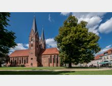 Backsteinkirche mit zwei hohen spitzen Türmen und roten Ziegeldächern, umgeben von einer Rasenfläche mit Bäumen. Rechts im Bild befinden sich Wohnhäuser und ein Café mit Sitzbereichen im Freien.