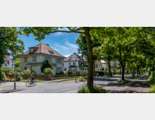 Wohnstraße mit Einfamilienhäusern im Stil der frühen 20. Jahrhundert-Architektur, umgeben von Grünflächen und hohen Bäumen. Im Vordergrund fahren Personen mit Fahrrädern auf einer Straße, die von einem Tempo-30-Schild markiert ist.