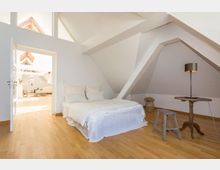 Schlafzimmer in einer Dachgeschosswohnung mit hellen Holzböden, weiß gestrichenen Wänden und schrägen Decken. Ein Bett mit weißer Bettwäsche steht links, daneben ein kleiner Holztisch mit einer Lampe und ein Hocker; im Hintergrund ist ein weiterer Raum mit sichtbaren Holzbalken erkennbar.