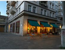 Ecke eines mehrstöckigen Wohnhauses mit heller Fassade, im Erdgeschoss ein Café mit großen Fenstern, grünen Markisen und warm beleuchteter Inneneinrichtung. Vor dem Café stehen bunte Tische und Stühle auf einem gepflasterten Platz.