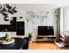 Moderne Wohnküche mit weißen Schränken, einer schwarzen Arbeitsfläche und einer eingebauten Mikrowelle und Backofen. Im Wohnzimmerbereich stehen ein großer Flachbildfernseher auf einem Holz-TV-Schrank, eine Ledersessel, ein schwarzer Esstisch mit Obstschale sowie eine grüne Pflanzenmuster-Tapete im Hintergrund.