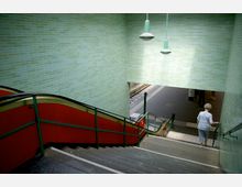 Eine Treppe führt zu einer U-Bahn-Station mit grün gekachelten Wänden, roten Seitenwänden und einem grünen Geländer. Im Hintergrund ist der Bahnsteig mit Schienen und einer wartenden Person sichtbar.